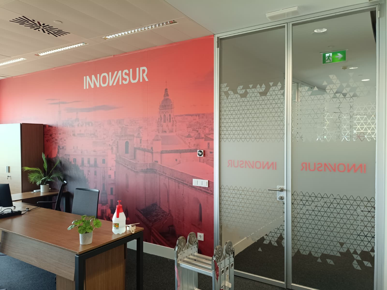 Montaje en las nueva oficina de “INNOVASUR” en Tore Sevilla, con mural rojo referencia “vista desde Mirador las Setas ”, puertas de vidrio con efecto ácido corporativo.